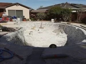 In-ground pool shell with white shotcrete finish, drain fitting, and stacked construction materials in a residential backyard