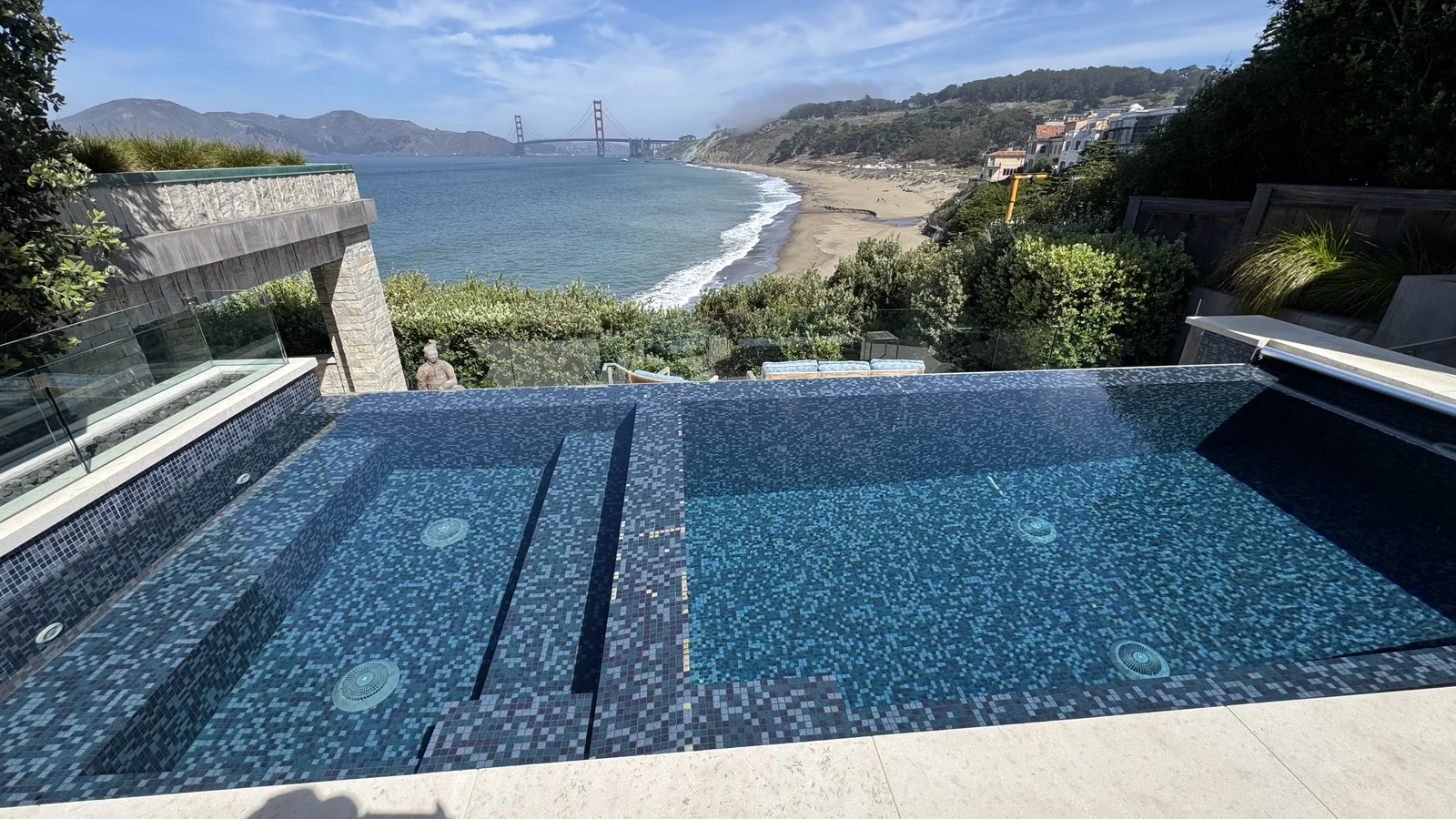 Infinity pool with blue and gray glass mosaic tile, stone coping, frameless glass railing, and recessed lighting