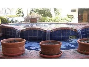 Blue glazed tile water feature with three cascading spouts over terracotta-toned ledge, brick pavers and terracotta planters in foreground