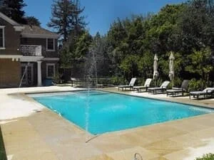 Rectangular in-ground pool with blue water, tan stone deck, fountain jet, and gray lounge chairs