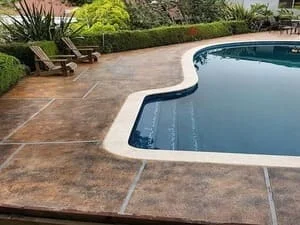 Freeform in-ground pool with beige coping and brown stamped concrete deck, lined by shrubs and wood lounge chairs