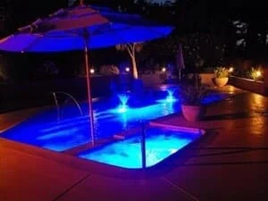 In-ground spa with blue underwater LED lighting beside a pool, tan textured concrete deck, and patio umbrella