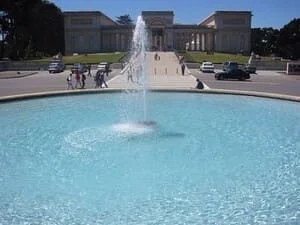 Round light-blue fountain basin with central water jet and paved plaza in front of a stone building