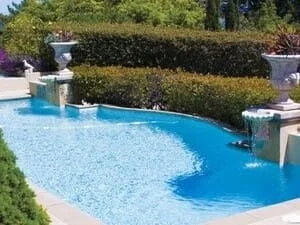 Freeform in-ground pool with blue water, stone urn planters, and spillway water features framed by a dense green hedge