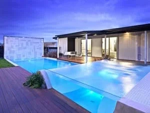 Illuminated in-ground pool with glass edge panel, dark timber deck, and modern glass-door house at dusk