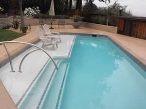 In-ground rectangular pool with light blue finish, tiled entry steps, stainless steel handrails, and tan concrete deck