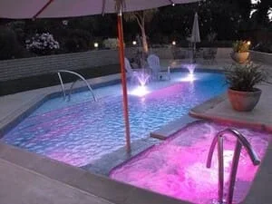 Night view of in-ground pool with pink and blue underwater LED lights, attached spa with stainless steel handrails, and patio umbrella