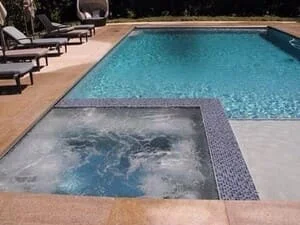 In-ground pool with light blue tile and raised spa with gray mosaic tile border on tan stone deck