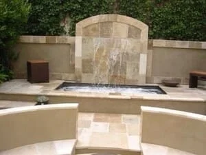 Arched travertine wall fountain with cascading water into a recessed basin, surrounded by beige travertine pavers and stone seat walls