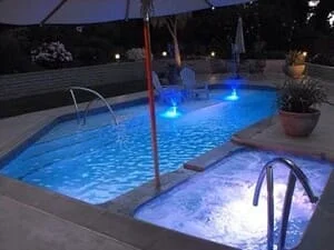 In-ground pool and attached spa with blue underwater lighting, stainless steel handrails, and light stone deck at night